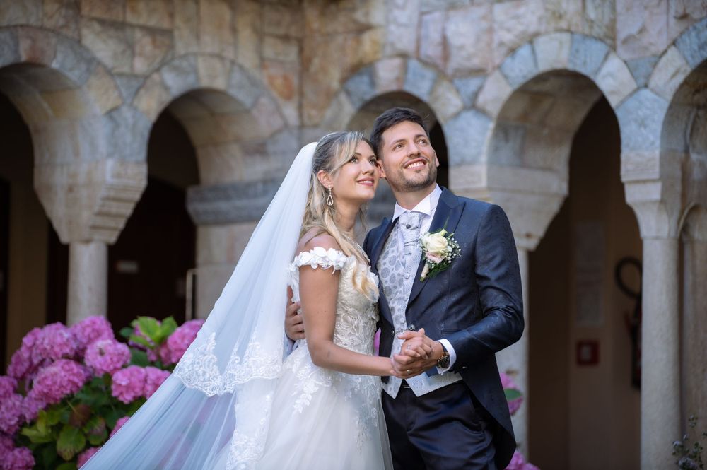 Elegant wedding couple poses in front of stone archways and pink hydrangeas at a historic venue.