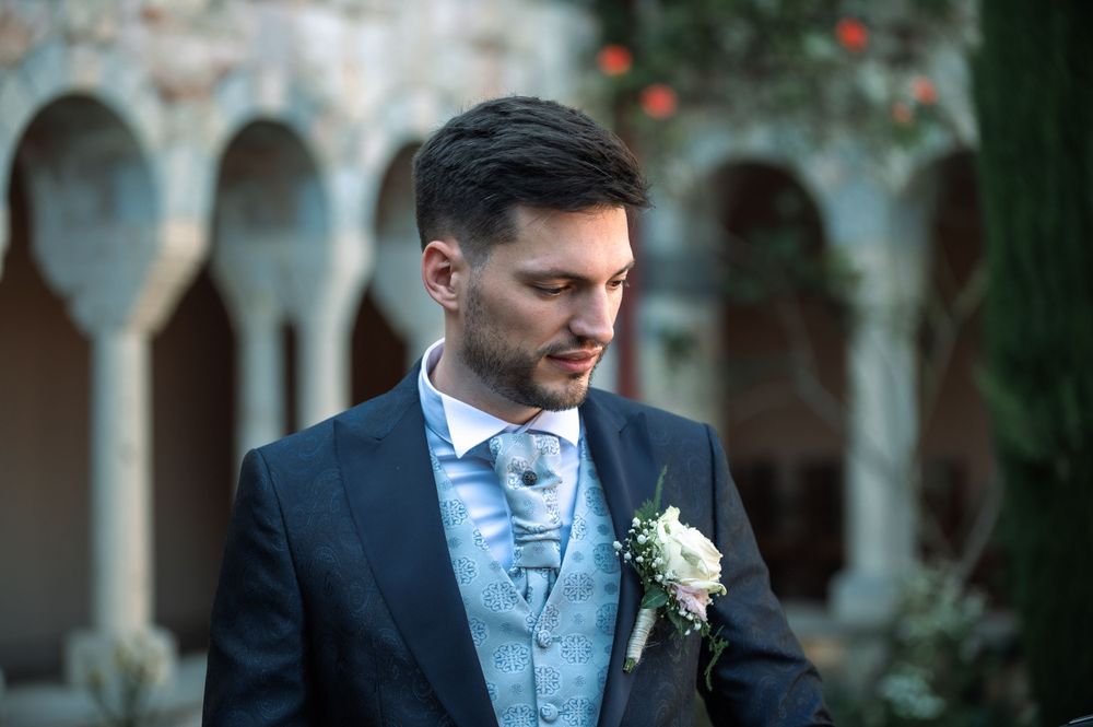 Groom in navy suit and light blue vest poses by stone colonnade.