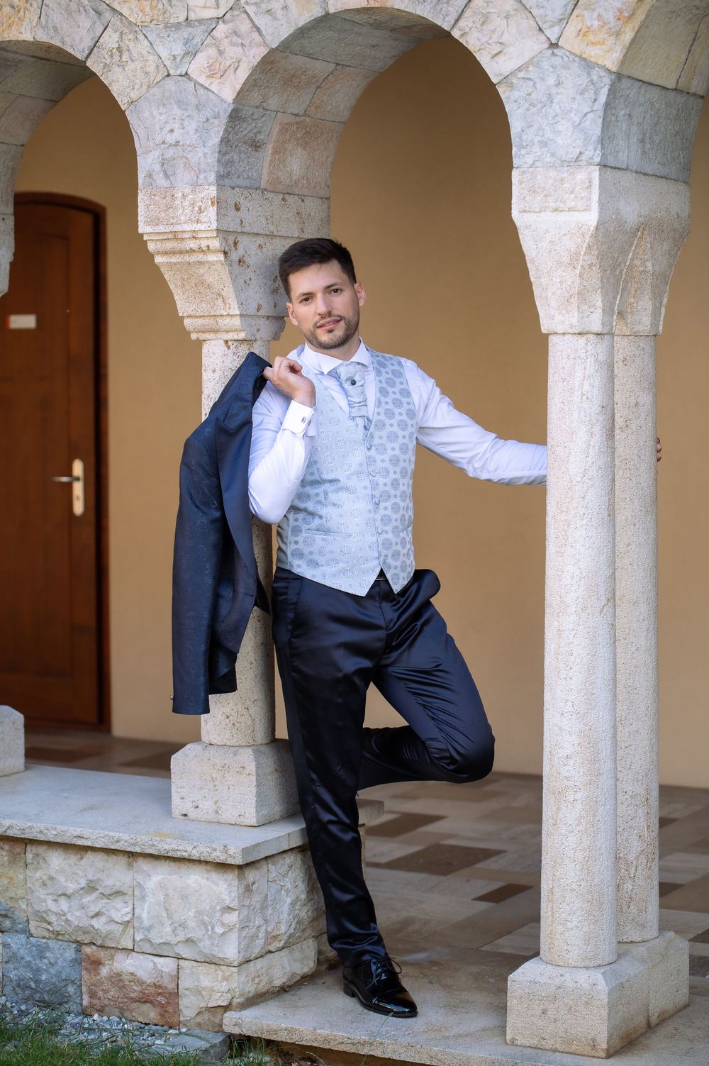 Playful groom poses between stone columns in formal wedding attire.