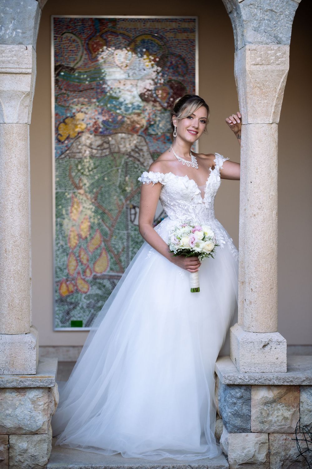 Bride in flowing tulle gown poses against weathered stone colonnade with painted wall murals.