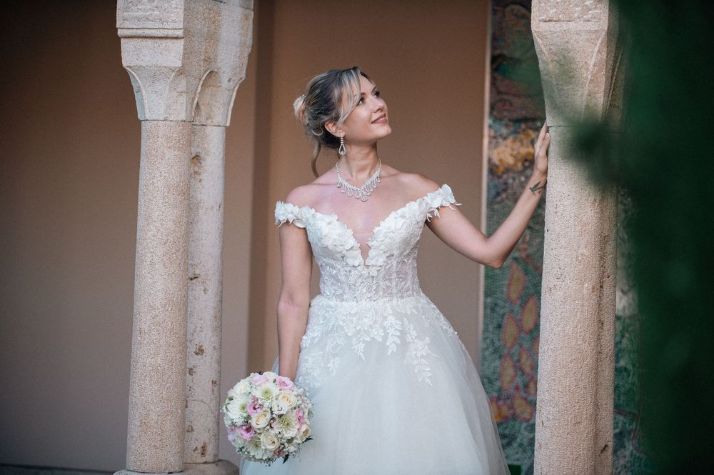 Bride in detailed lace gown poses under historic stone archways.