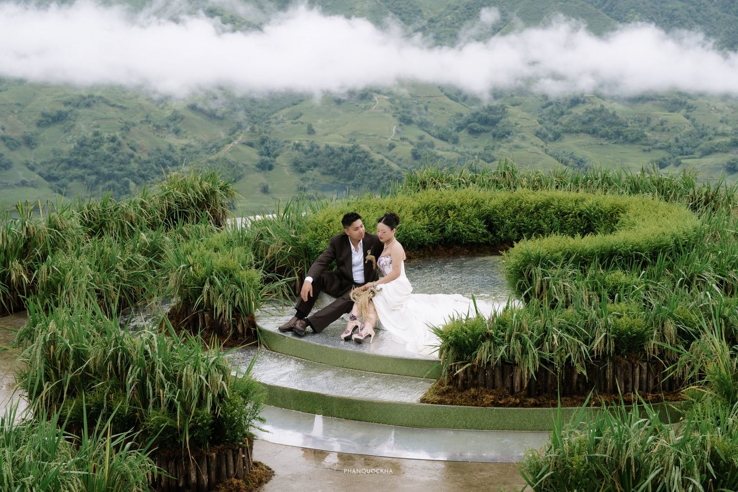 Bride and groom sit together on curved terraced rice field path surrounded by green mountain scenery.