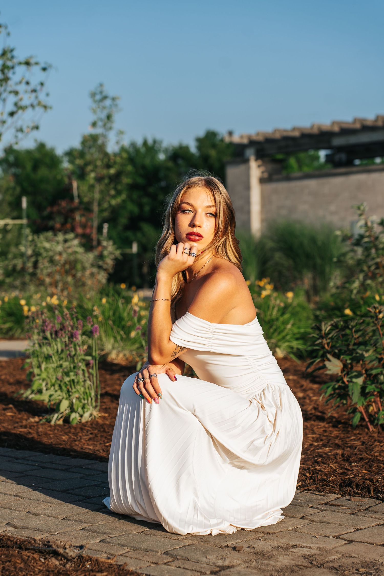 Woman in white off-shoulder dress poses in garden setting during golden hour.