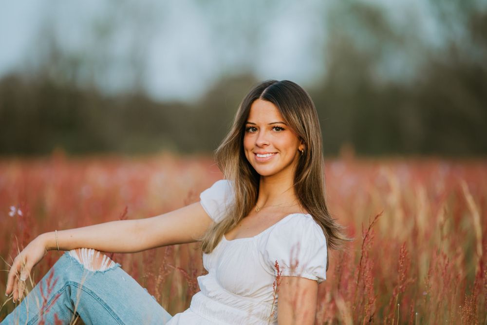 A young woman in a white top sits in a golden wheat field during sunset with a warm glowing background.