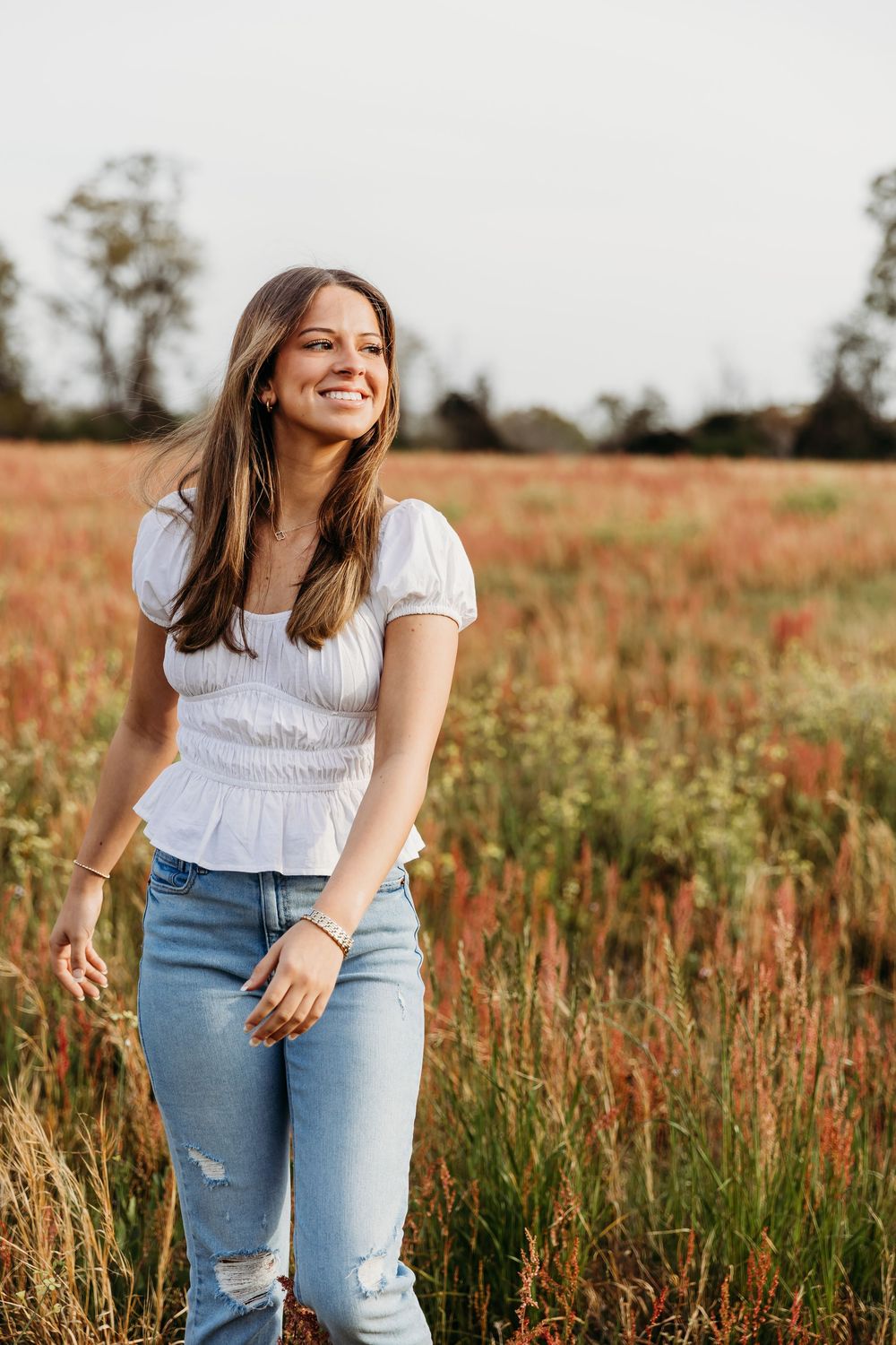 A girl in a white smocked top and ripped jeans stands smiling in a golden summer field at sunset.