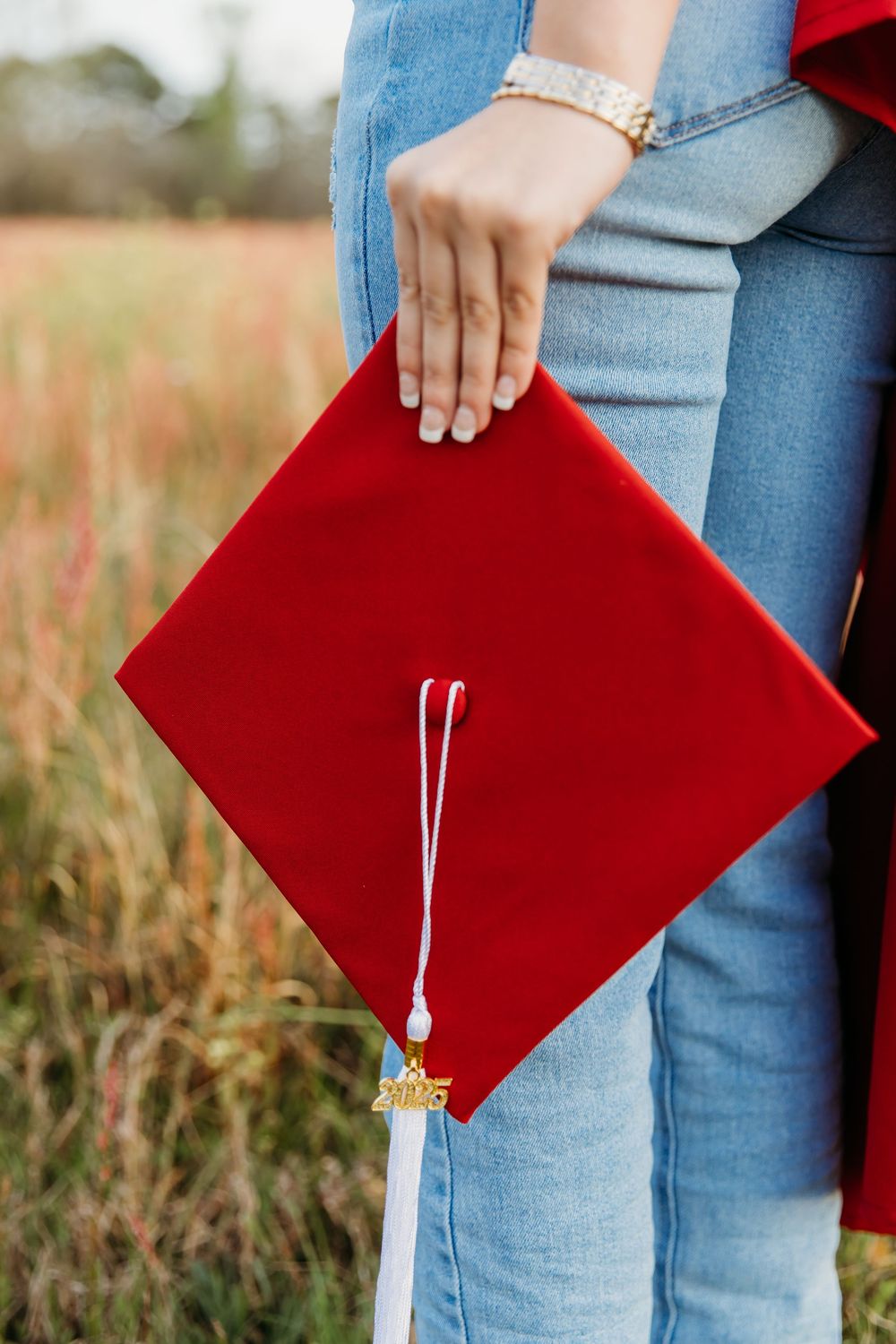 A hand holding a red graduation cap with tassel against a blurred natural background.