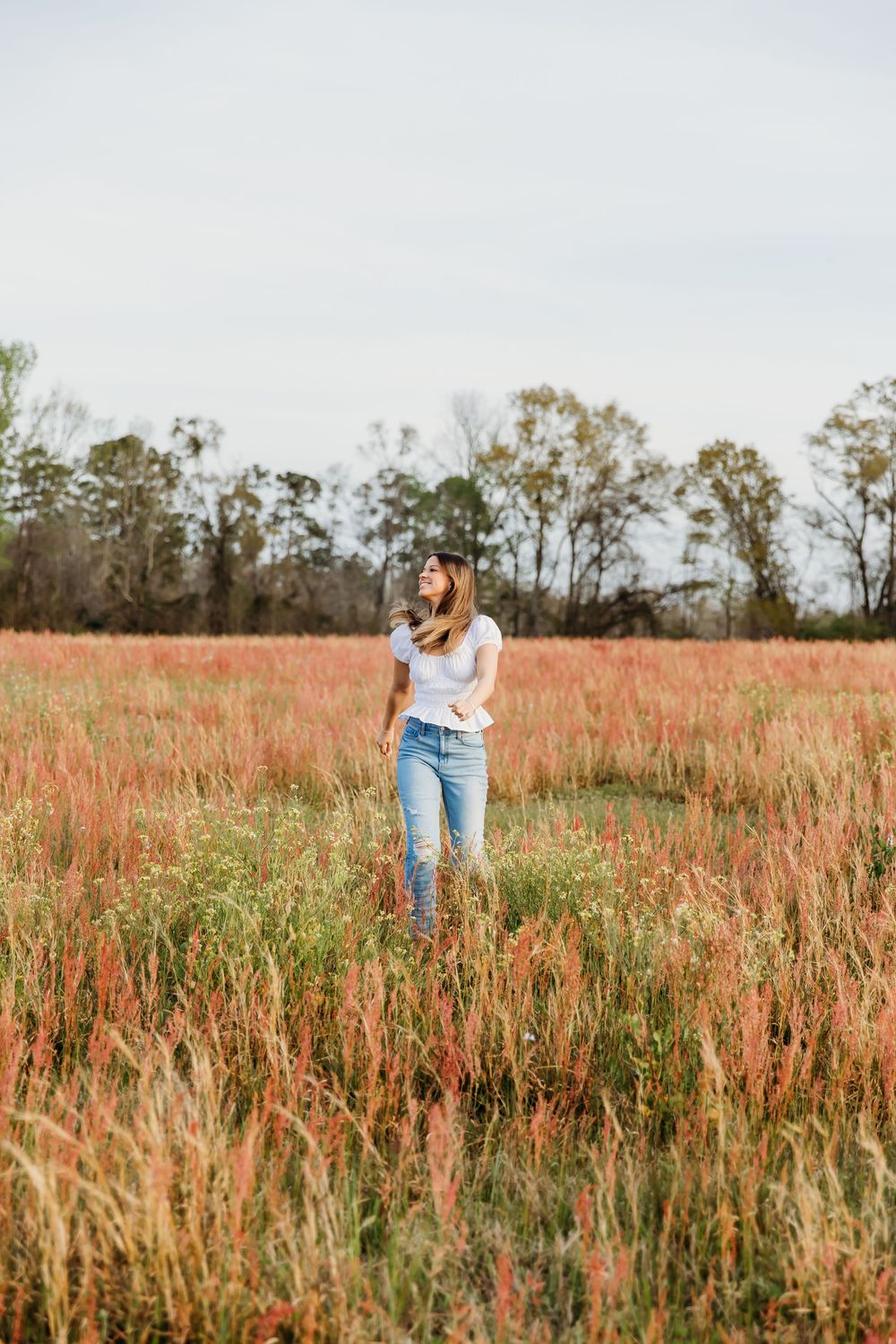 A woman stands in a rustic field of tall reddish grass and wildflowers during golden hour.