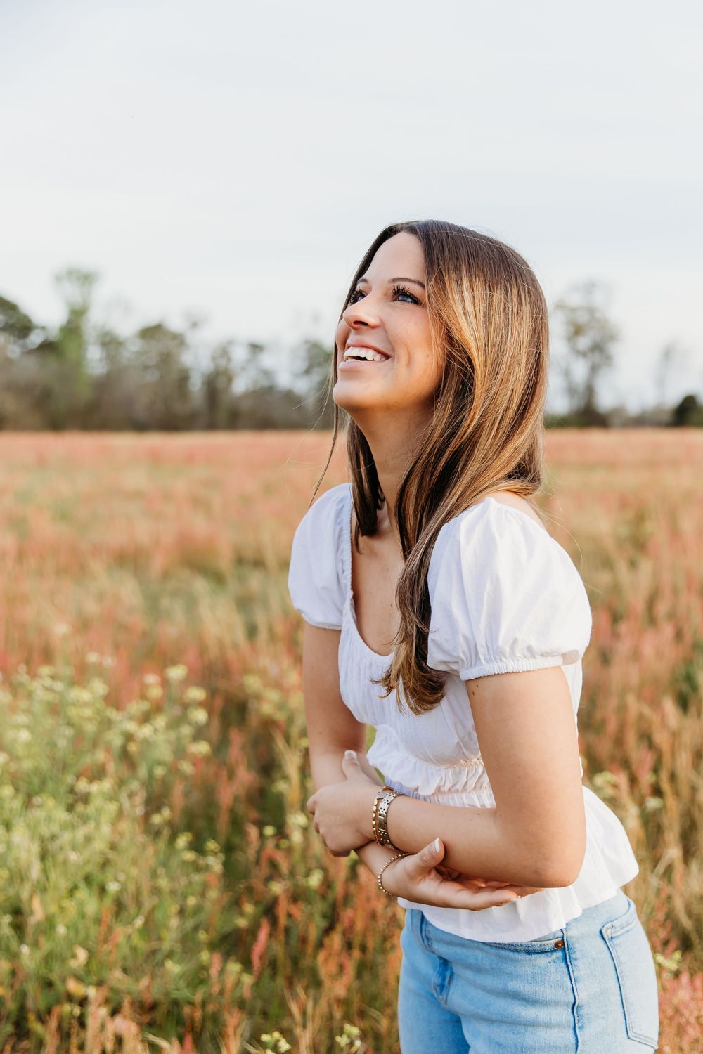 Person in a white top and light jeans laughs joyfully in a wildflower field during golden hour.