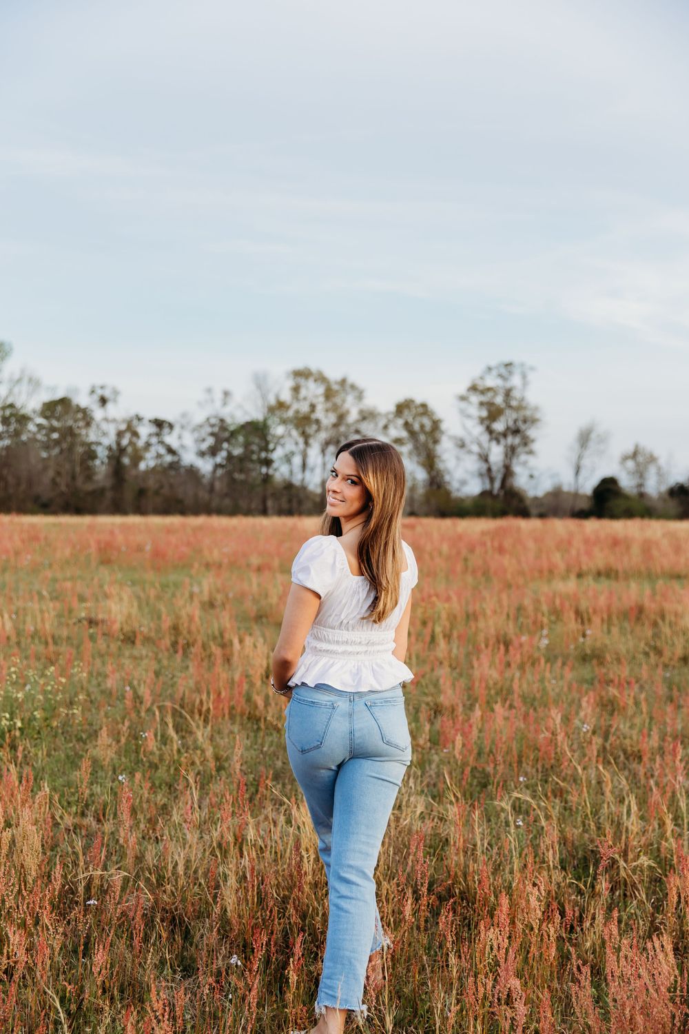 A person in a white top and jeans stands amongst tall reddish-brown grass in a golden field during sunset.
