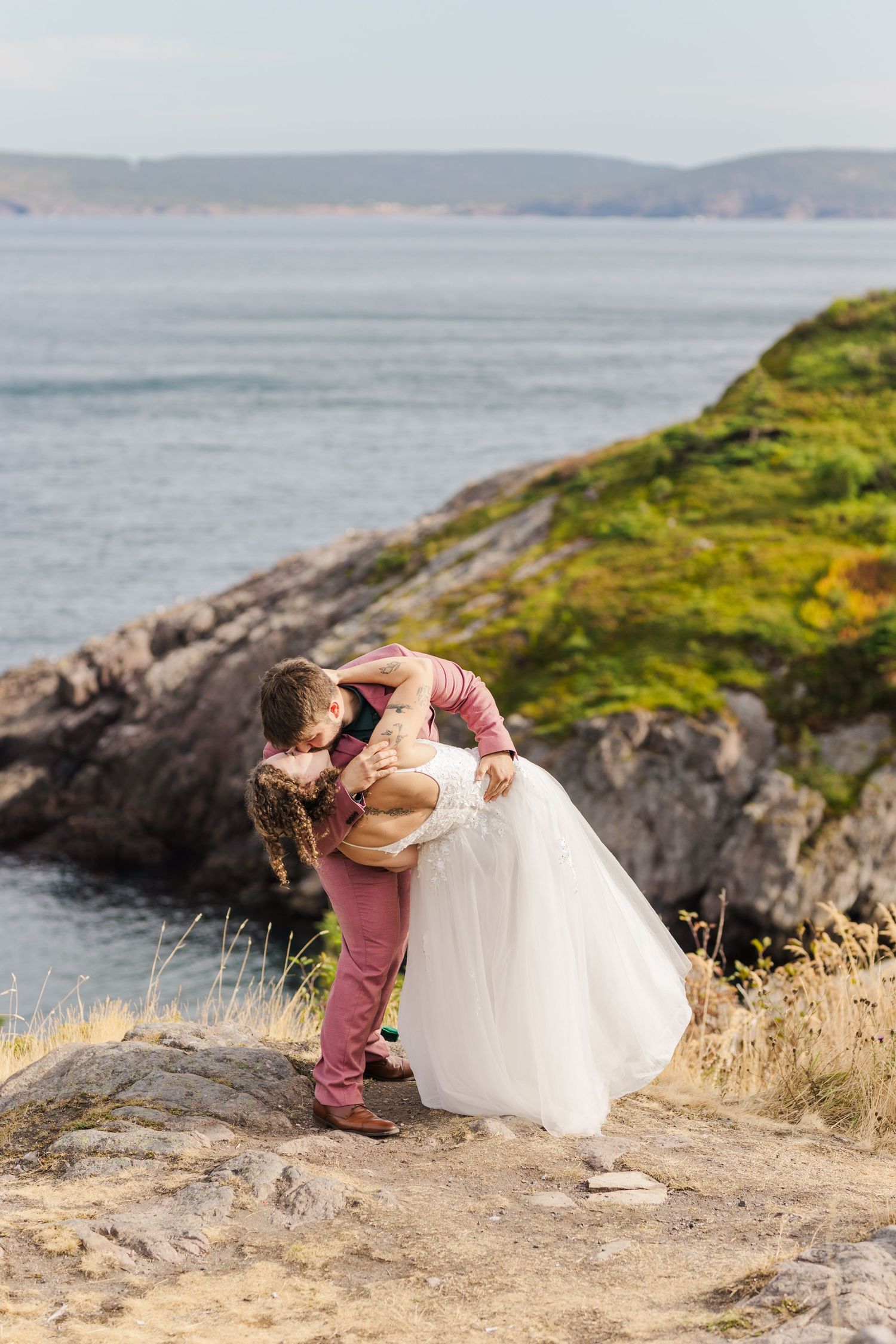 A couple shares a romantic moment on a coastal cliff overlooking the ocean during sunset.