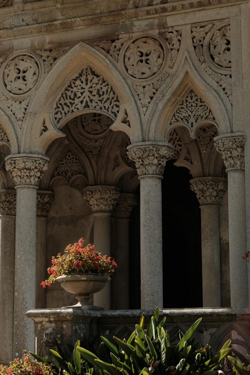 Ornate stone columns and arches in a monastery cloister with flowering plants and sunlight streaming through the arcade.