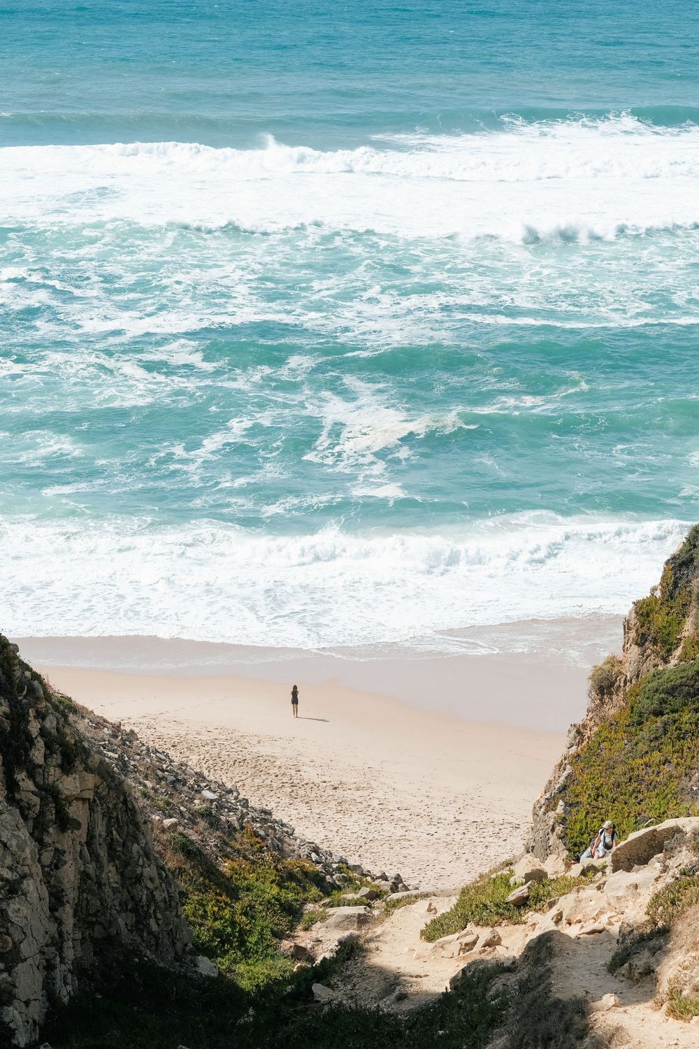 Aerial view of rugged coastal cliffs overlooking turquoise ocean waves crashing on a sandy beach with a lone figure below.