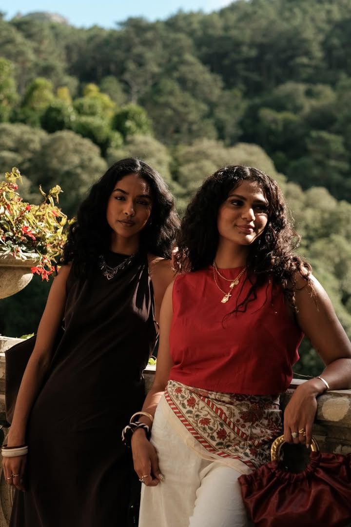 Two women in contrasting black and red outfits pose against a scenic forest backdrop on a sunny afternoon.