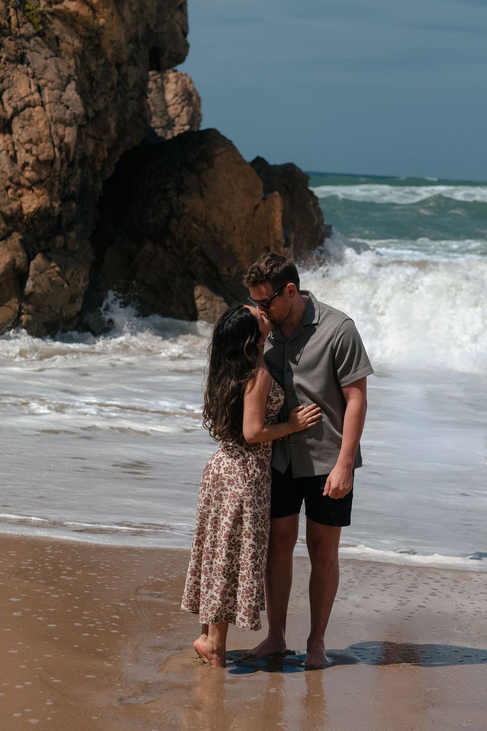 A couple shares an intimate moment on a beach with crashing waves and rocky cliffs in the background.