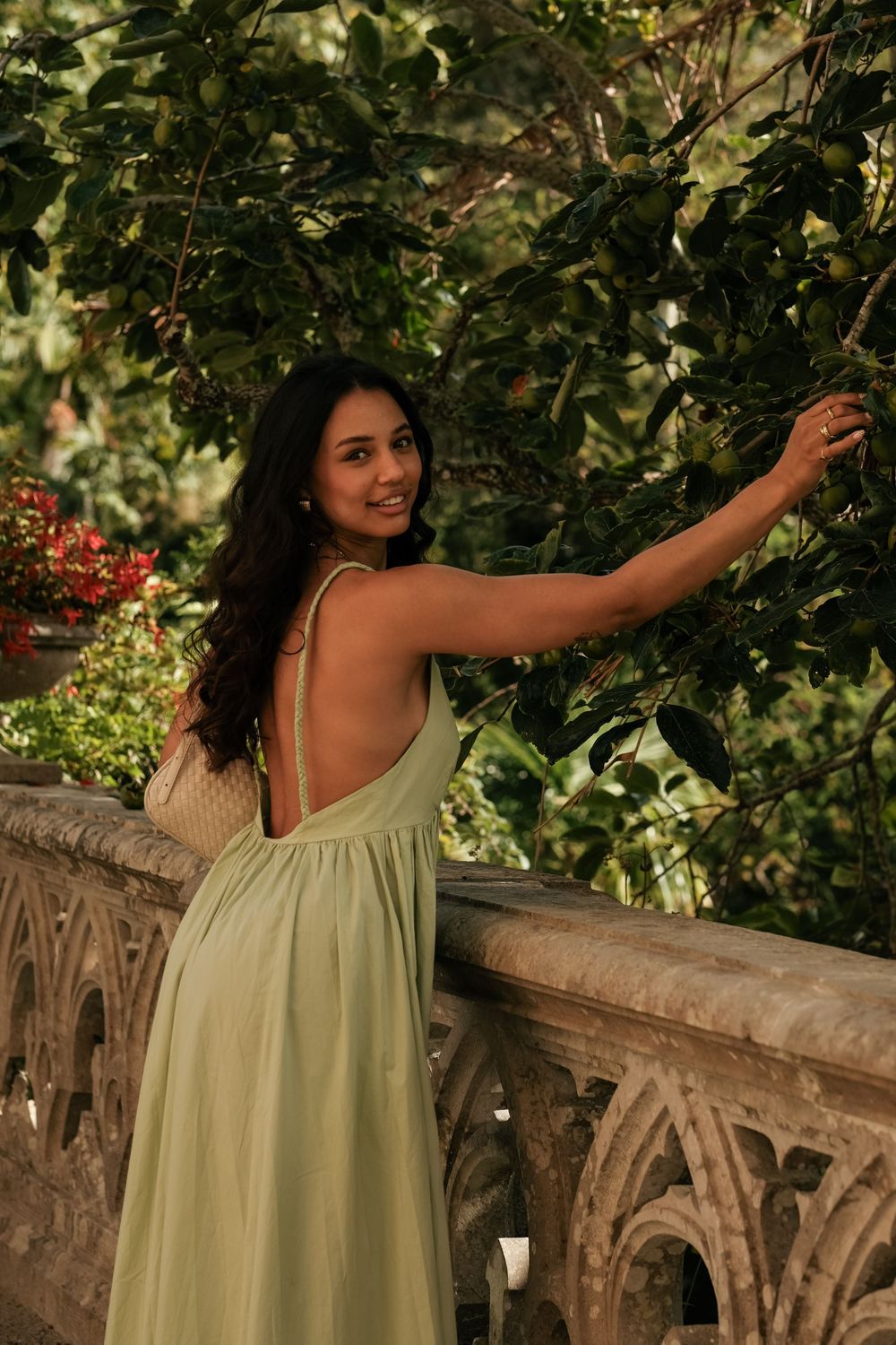 A woman in a light green dress poses against a stone railing surrounded by lush greenery in a garden setting.