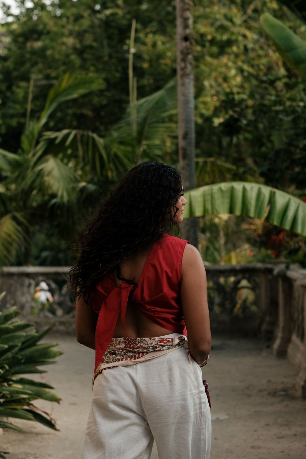 Someone in a red top and white skirt walking through a lush tropical garden path with palm trees and greenery.