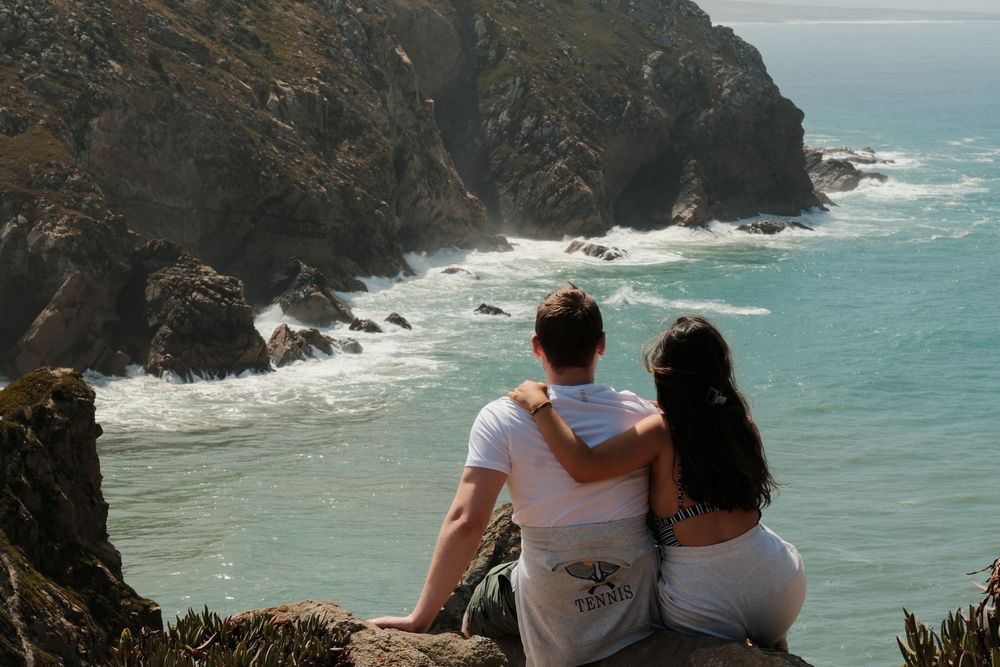 People sitting at the edge of a rocky cliff overlooking the turquoise ocean and rugged coastline below.