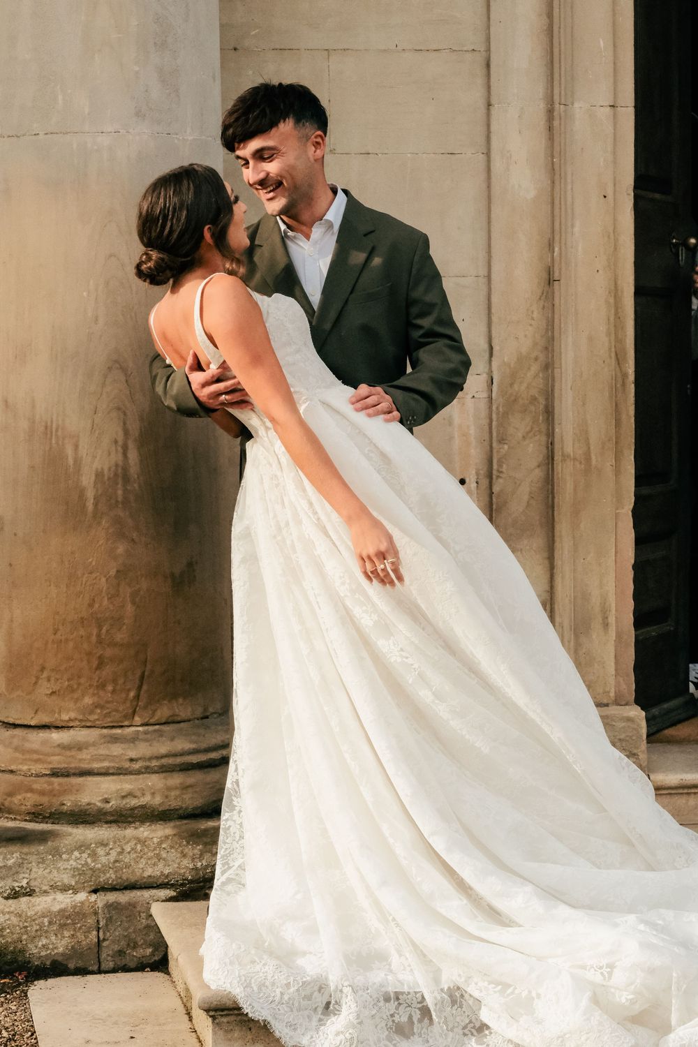 A romantic wedding couple embraces and laughs together while standing by a stone column, bride wearing a flowing lace gown.