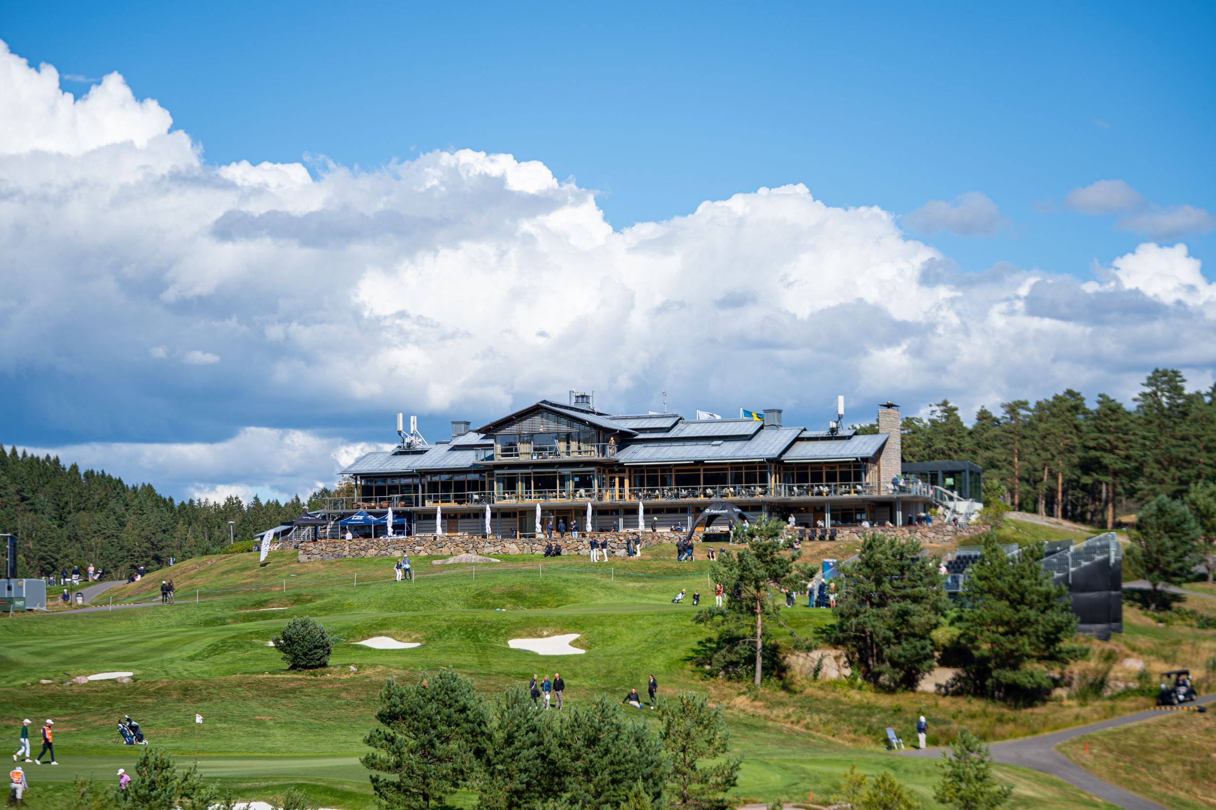 A scenic golf course features a modern clubhouse nestled among rolling green hills against a bright blue sky.