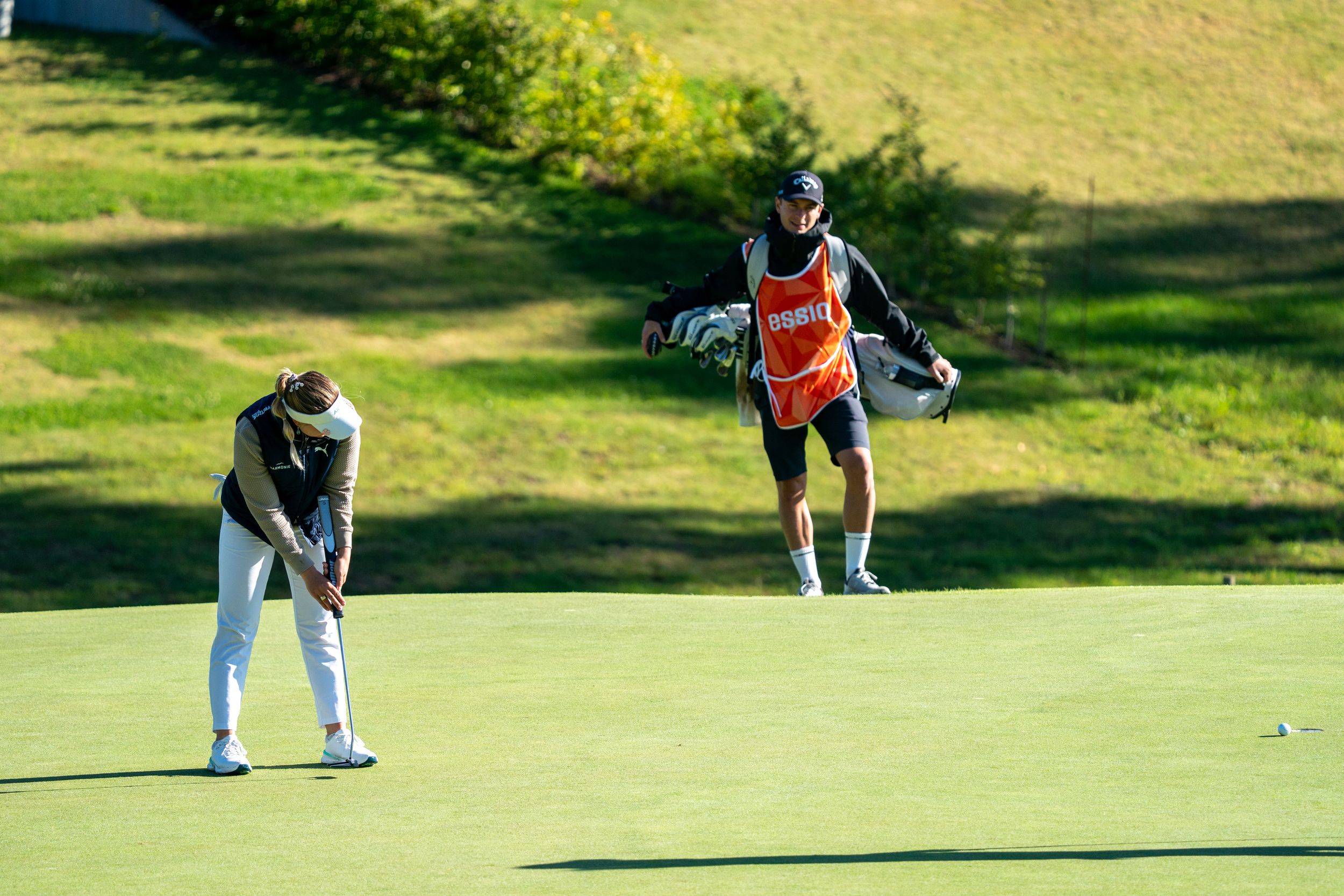 Golfer in orange shirt lines up putt on sunny green while others watch on golf course.