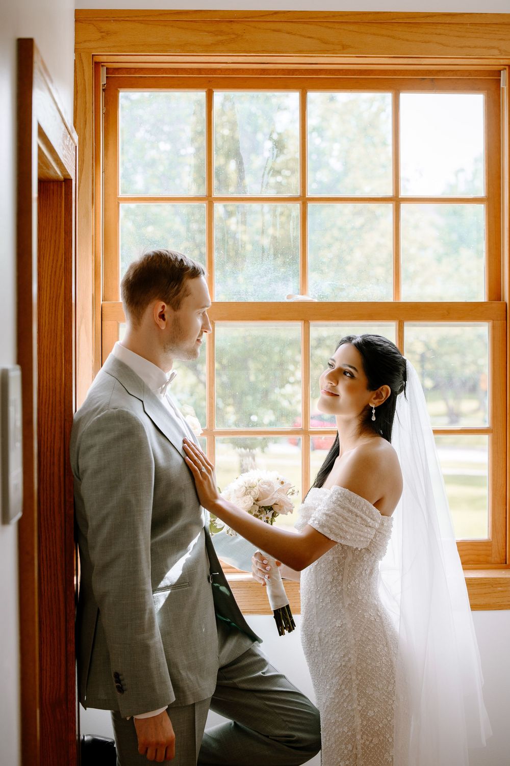 Tender moment between newlyweds standing by a sunlit window as the bride adjusts the groom's tie on their wedding day.