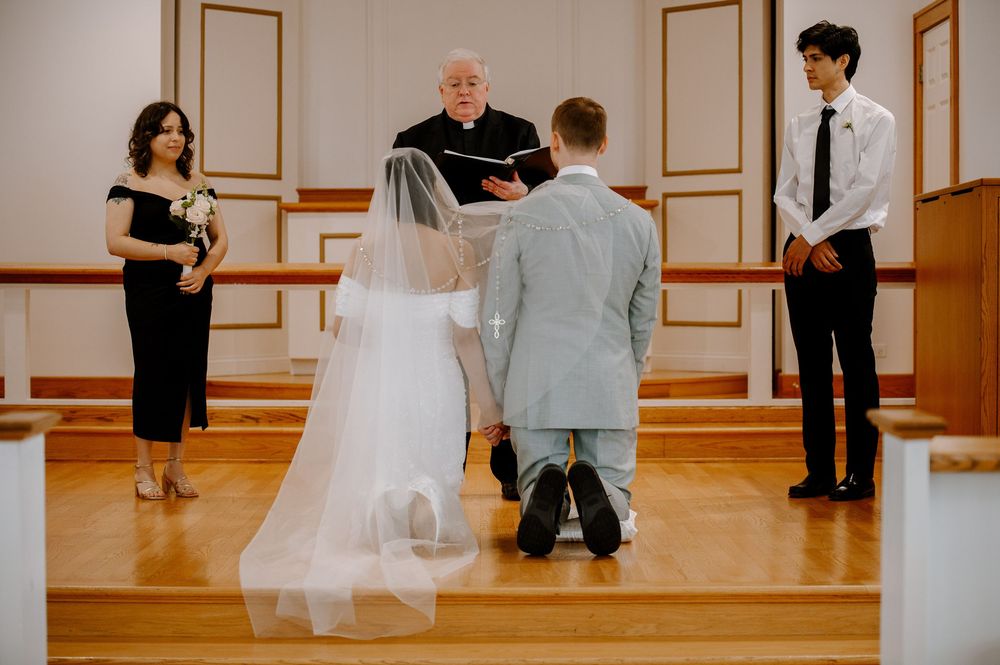 A wedding ceremony takes place in a chapel with wood floors and cream colored walls as a couple kneels at the altar.