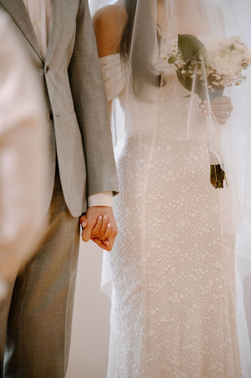 A close-up of a bride and groom holding hands, showing the bride's white lace dress and groom's gray suit.
