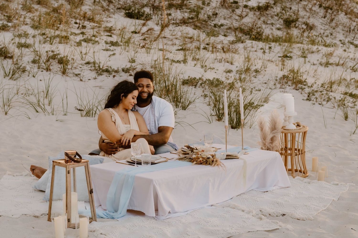 Romantic beach picnic setup with white linens and decorative elements.