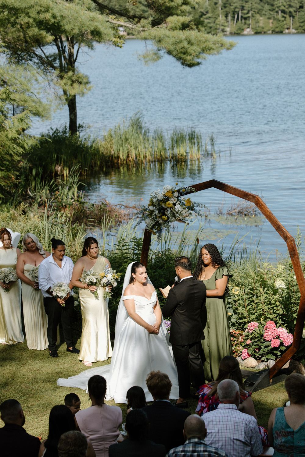 Outdoor lakeside wedding ceremony with bride and groom at floral arch, bridesmaids in sage green gowns.
