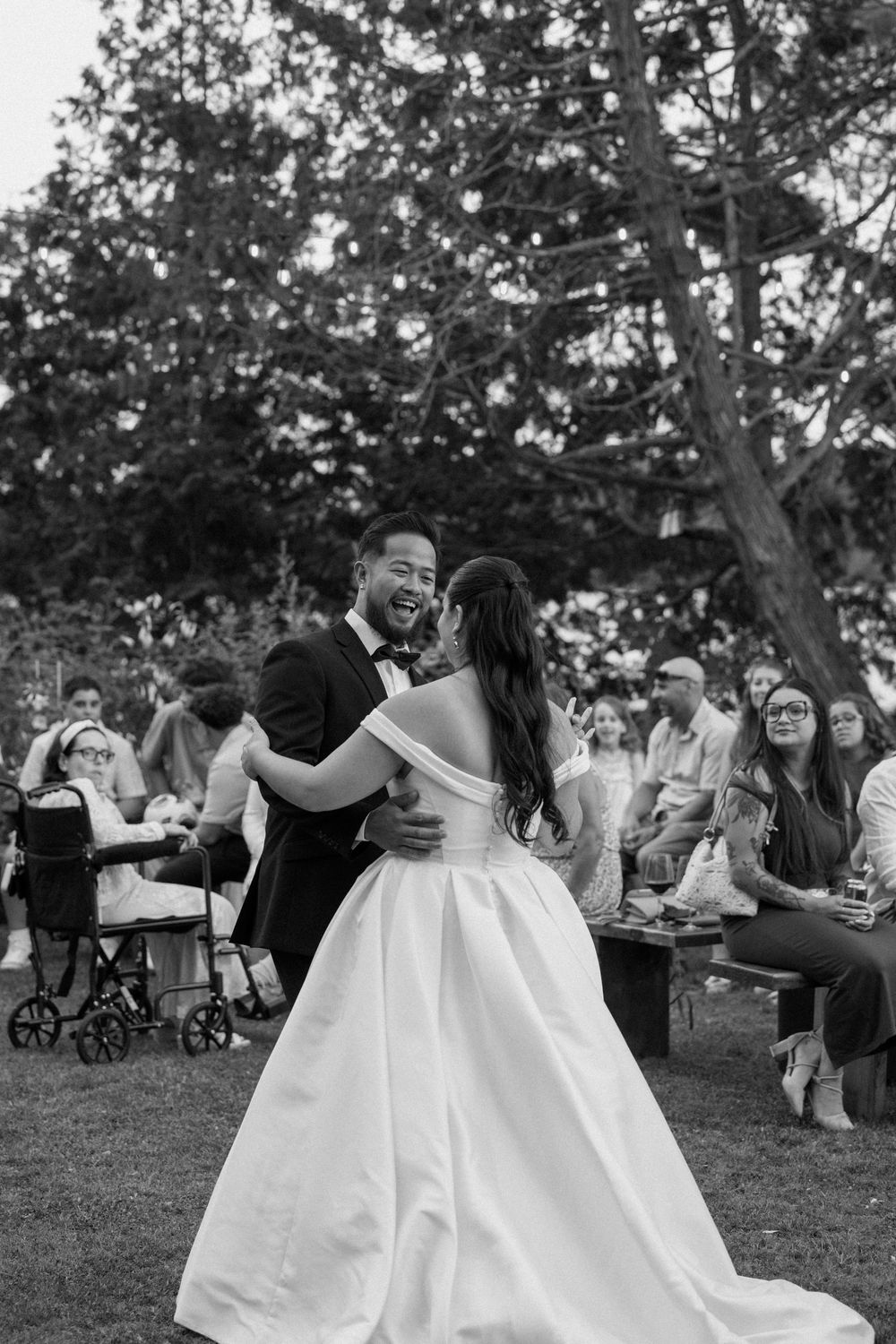 Black and white photo of bride and groom sharing first dance outdoors with guests seated around them.