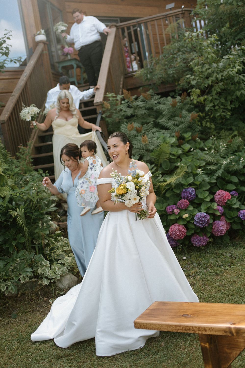 Smiling bride in white ball gown carries a colorful bouquet as her wedding party descends garden steps behind her.