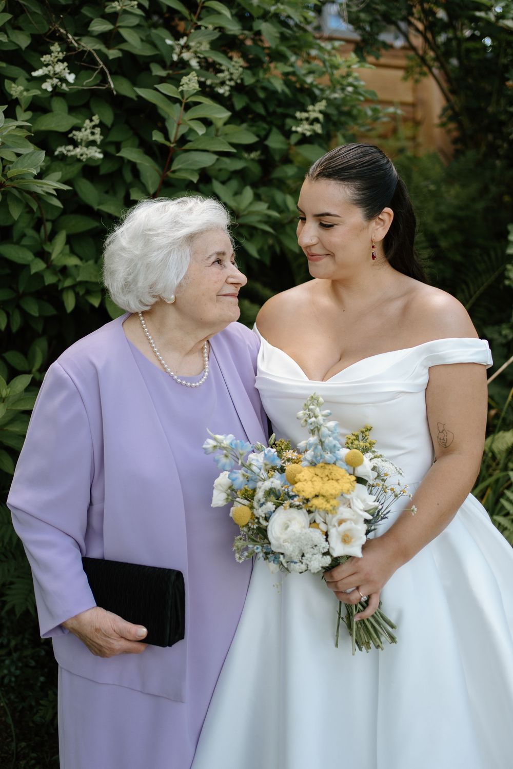 Bride in white off-shoulder gown holding a yellow and blue bouquet shares a warm smile with an elderly woman in lavender.