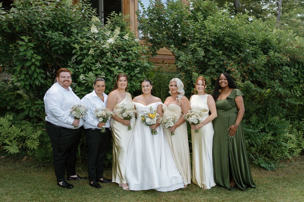 Bride with bridesmaids in ivory and green gowns holding white and yellow bouquets outdoors at wedding.