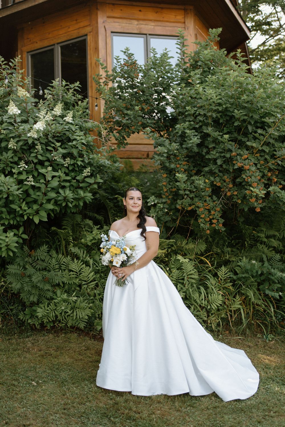 Bride in off-shoulder white ball gown holding yellow and white bouquet standing before rustic wooden cabin.