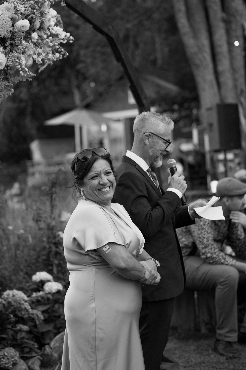 Black and white photo of smiling woman in cape dress with man giving speech at outdoor wedding.