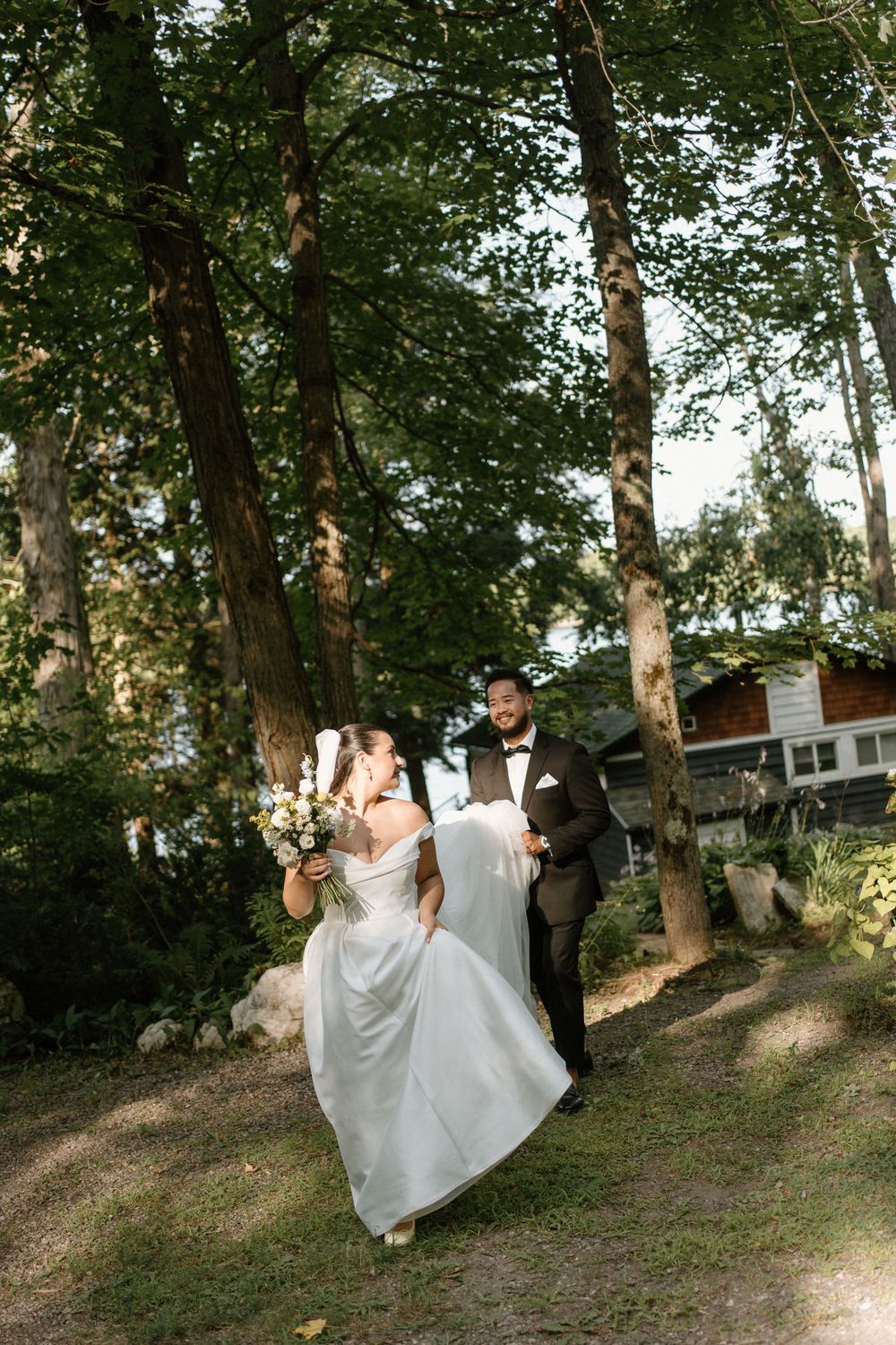 Bride and groom walk hand-in-hand through a sunlit forest path, bride holding a floral bouquet in her flowing white gown.