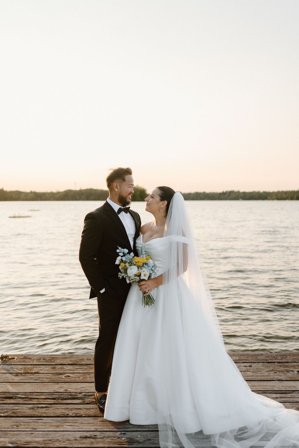 Bride and groom share a romantic moment on a wooden dock at sunset, bride holding a blue and yellow bouquet.