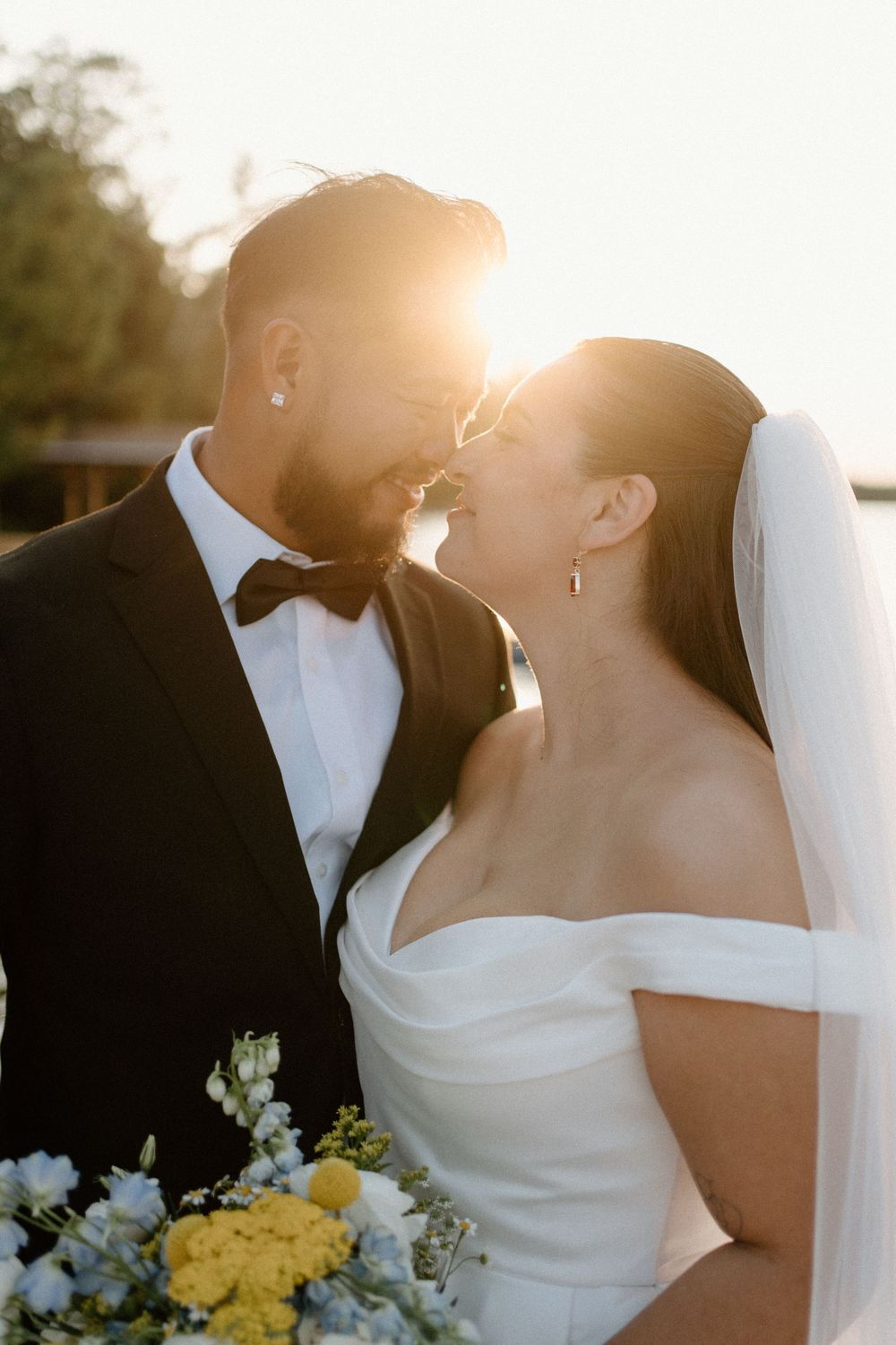 Bride and groom lean in for a kiss bathed in golden sunset light, bride holding a colorful floral bouquet.