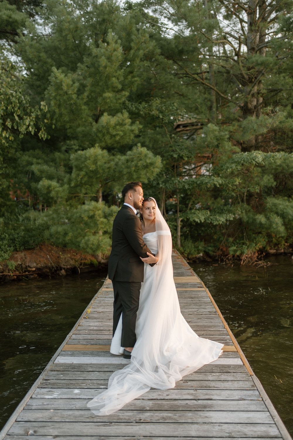 Bride and groom embrace tenderly on a wooden dock surrounded by green pine trees reflected in calm water.