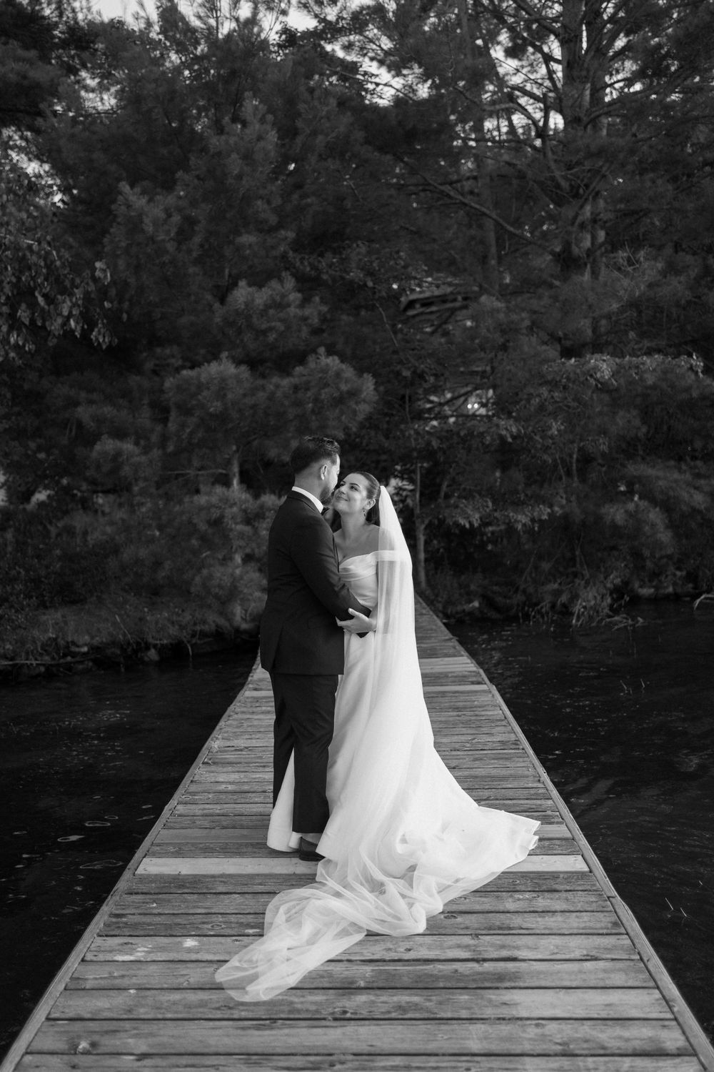 Black and white photo of bride and groom sharing a kiss on a wooden dock with pine trees behind them.