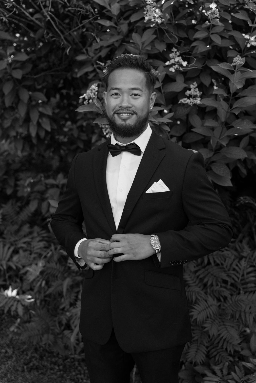 Black and white portrait of smiling groom in tuxedo with bow tie and pocket square outdoors.
