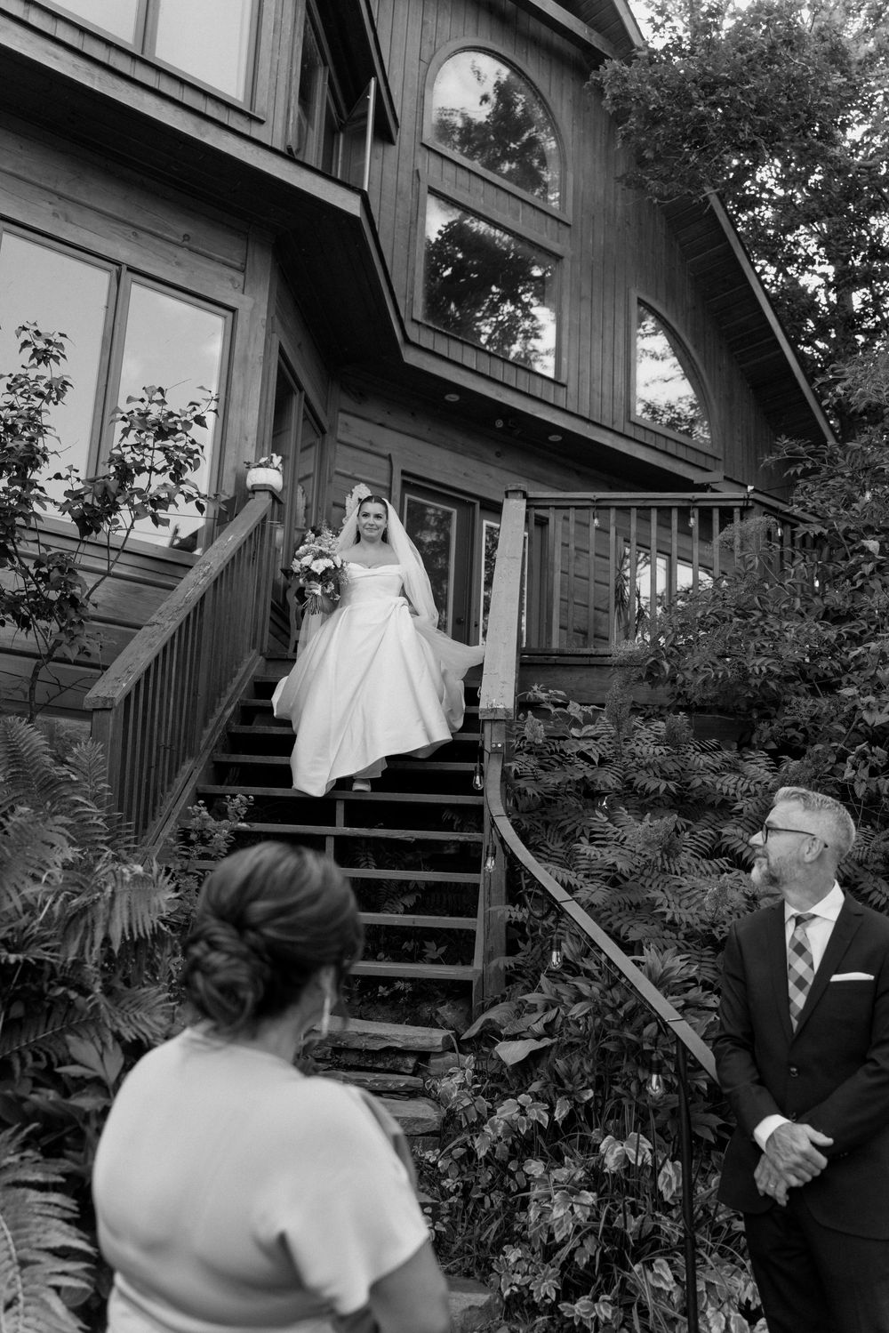 Black and white photo of bride in white gown descending outdoor staircase holding bouquet at venue.