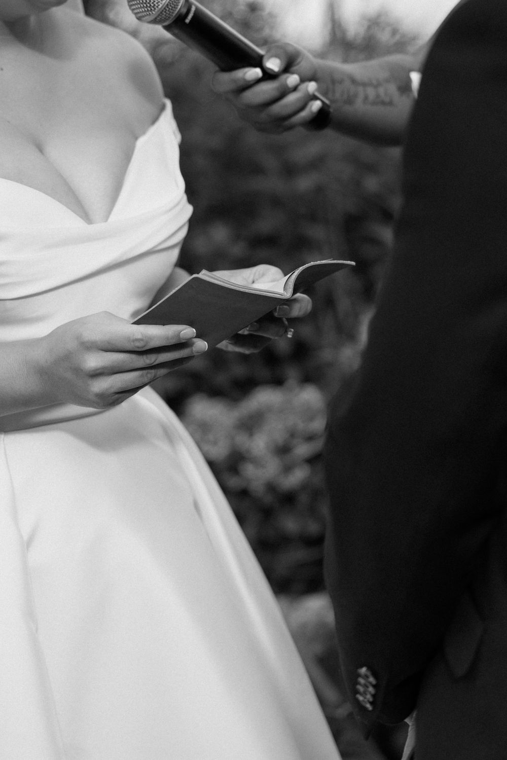 Black and white close-up of bride in strapless gown reading wedding vows from small booklet.