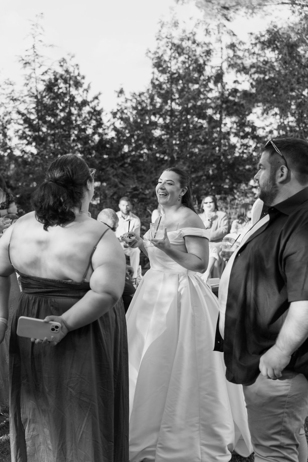 Black and white photo of smiling bride in off-shoulder gown mingling with wedding guests outdoors.