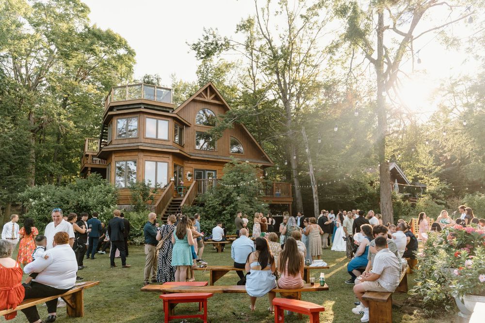 Outdoor wedding reception at a rustic wooden venue surrounded by trees with guests mingling on a sunny afternoon.
