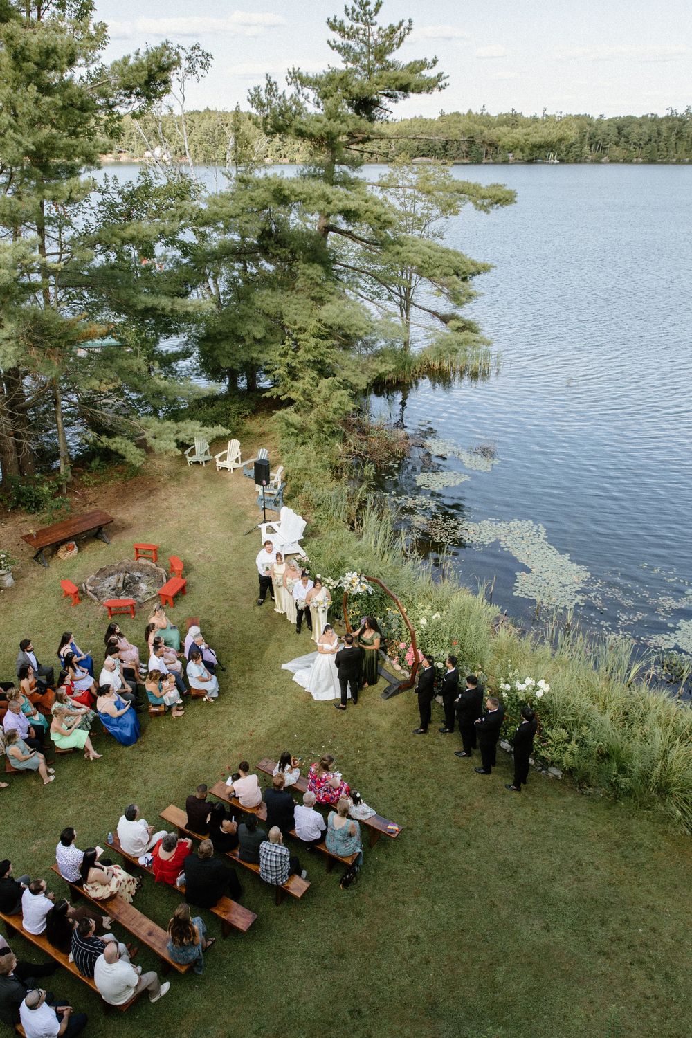 Aerial view of outdoor lakeside wedding ceremony with guests seated on wooden benches among pine trees.