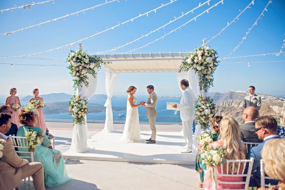 Decorative string lights and floral arrangements line a beachfront wedding ceremony setup against a bright blue sky and ocean.