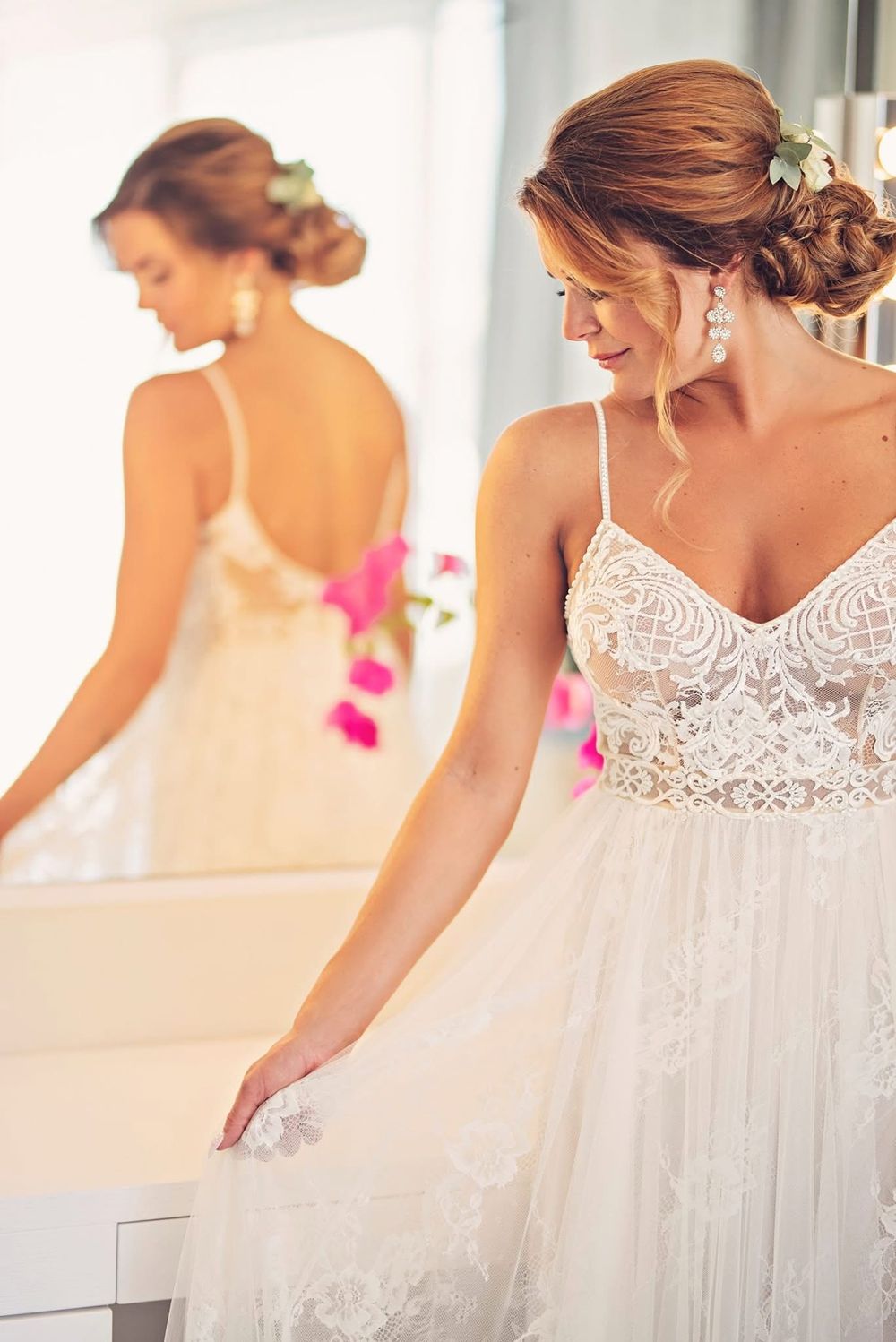 A bride in a delicate lace and tulle wedding gown admires herself in a mirror while wearing elegant earrings.