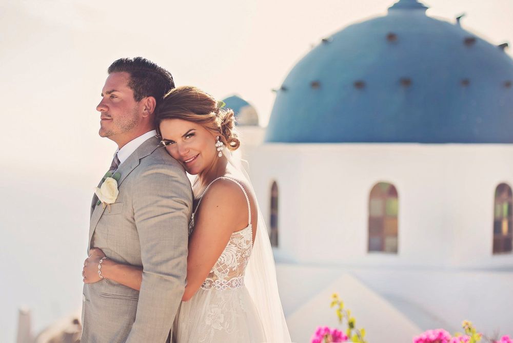 A newlywed couple shares romantic moments against a backdrop of white Mediterranean architecture and vibrant flowers.