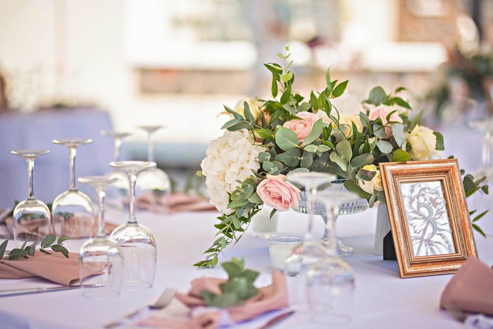Elegant wedding reception table setting with pink and white floral centerpiece, wine glasses, and framed photo decor.